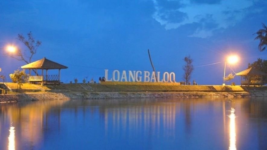 Lakeside park at dusk with a large sign reading 'LOANG BALOO', illuminated by street lamps and flanked by two gazebos overlooking calm water.