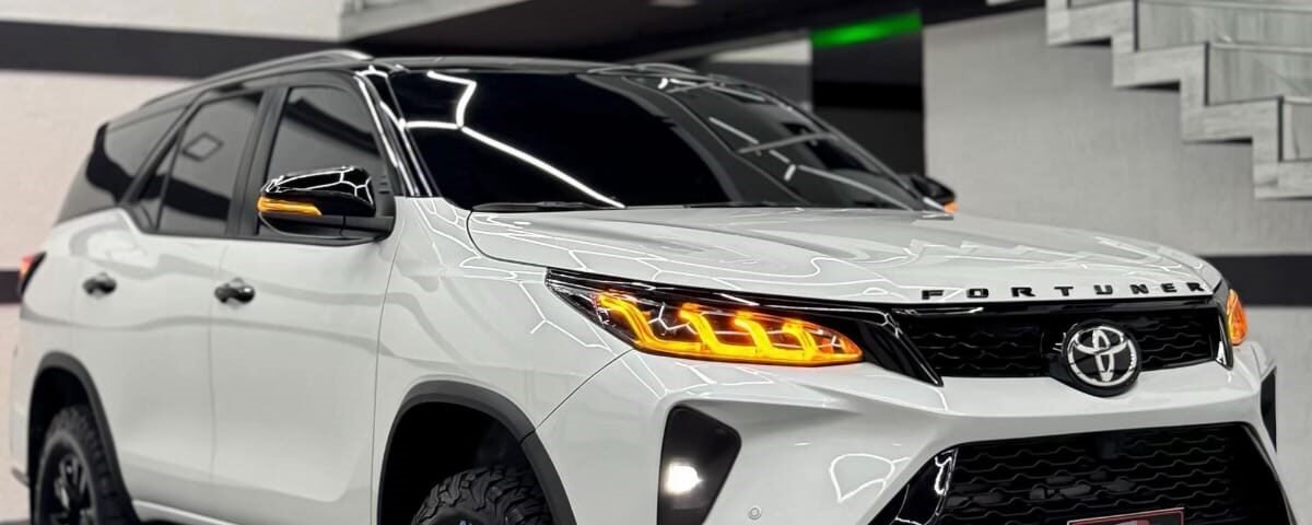 Front three-quarter view of a white Toyota Fortuner SUV in a modern showroom with glass railing and geometric ceiling lights in the background.