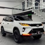 Front three-quarter view of a white Toyota Fortuner SUV in a modern showroom with glass railing and geometric ceiling lights in the background.