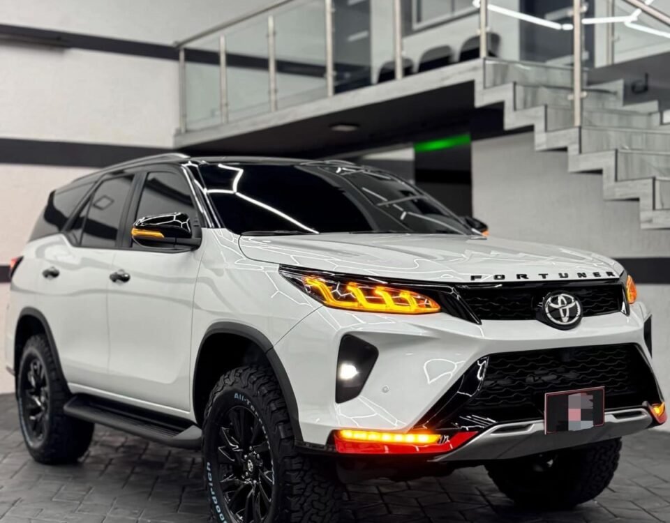 Front three-quarter view of a white Toyota Fortuner SUV in a modern showroom with glass railing and geometric ceiling lights in the background.