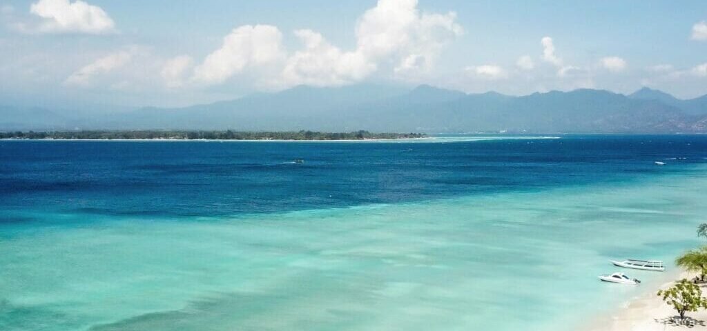 Turquoise shallow water along a white-sand beach with small boats and distant mountains under a blue sky.