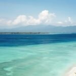 Turquoise shallow water along a white-sand beach with small boats and distant mountains under a blue sky.