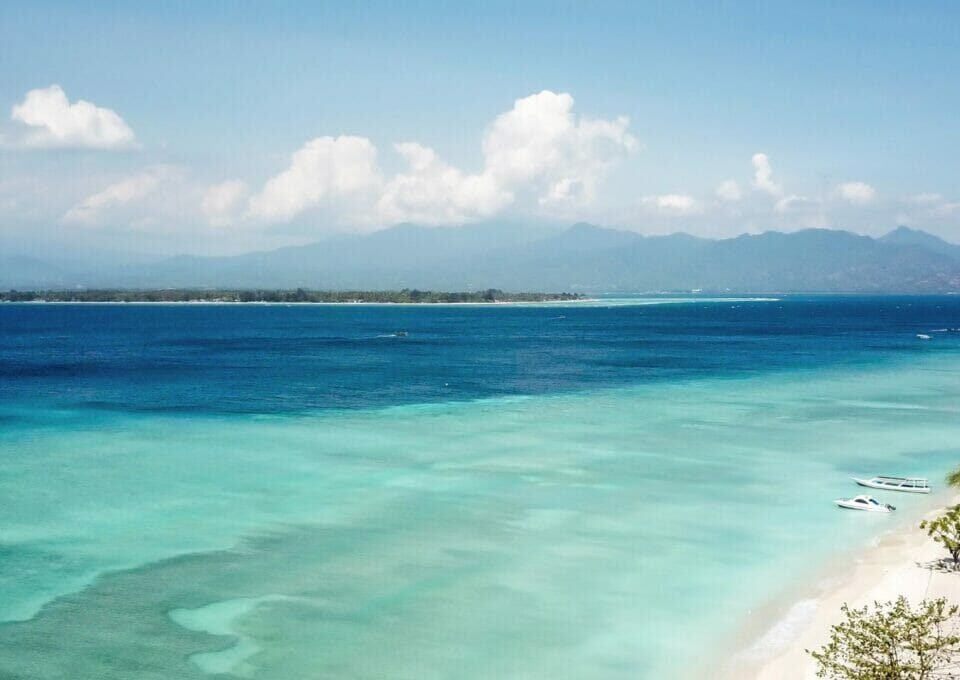 Turquoise shallow water along a white-sand beach with small boats and distant mountains under a blue sky.