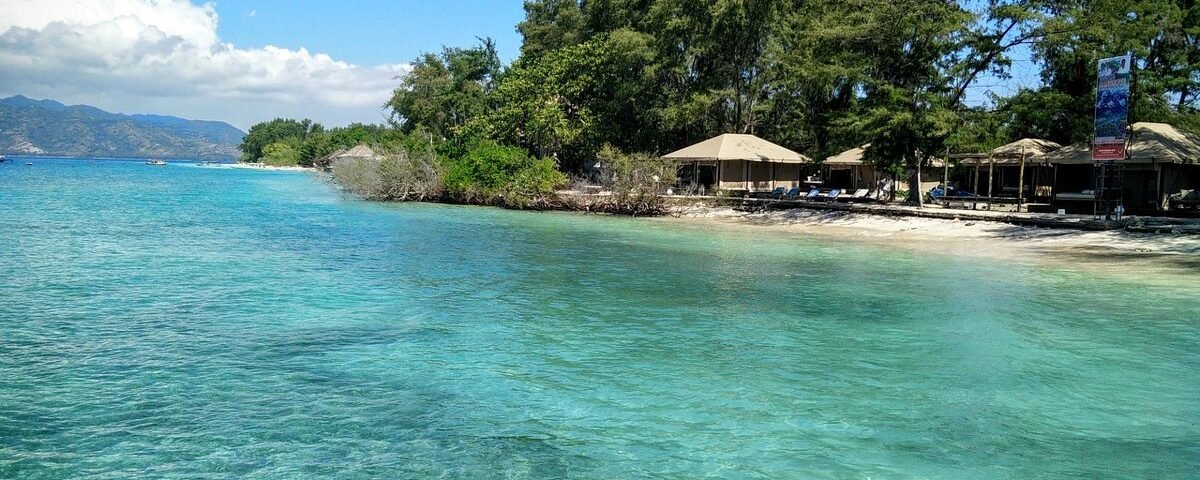 Turquoise water and a sandy beach lined with thatched umbrellas and trees along a sunny coastline.