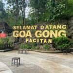Large yellow sign reading 'Selamat Datang Goa Gong Pacitan' at the entrance of a rocky hillside park, with string lights, trees, and benches in the foreground.