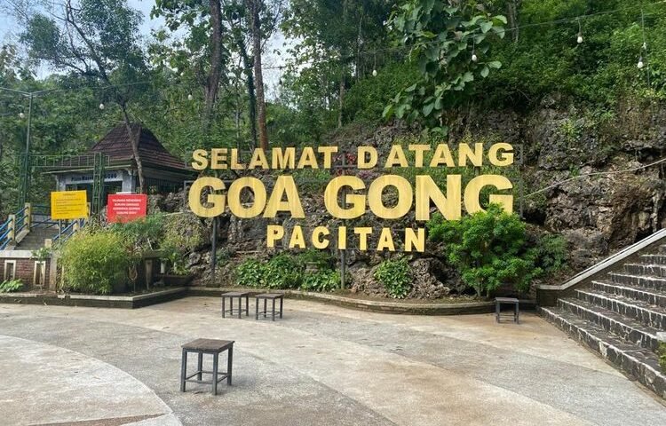 Large yellow sign reading 'Selamat Datang Goa Gong Pacitan' at the entrance of a rocky hillside park, with string lights, trees, and benches in the foreground.