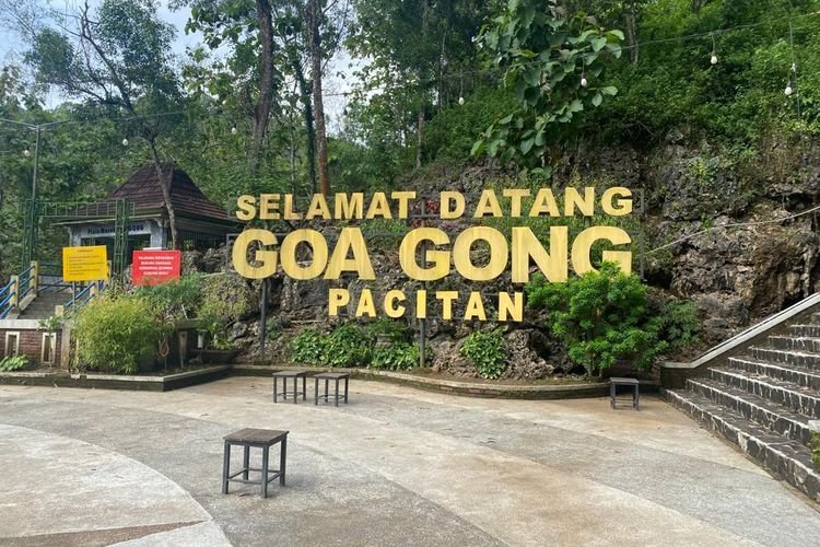Large yellow sign reading 'Selamat Datang Goa Gong Pacitan' at the entrance of a rocky hillside park, with string lights, trees, and benches in the foreground.