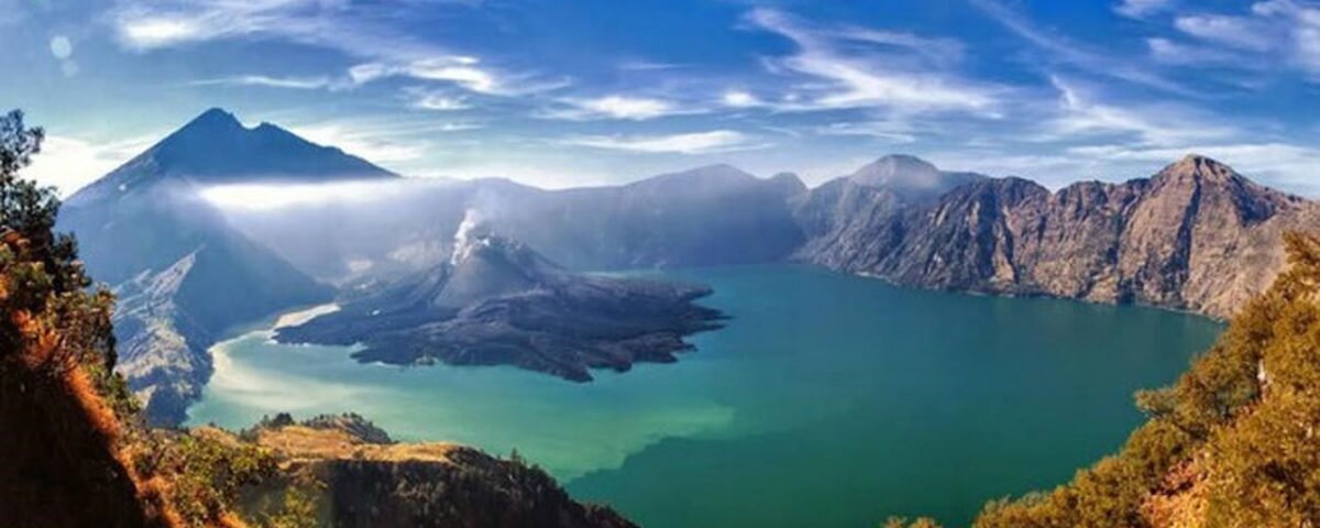 Panoramic view of a caldera lake with turquoise water, surrounded by rugged mountains and a blue sky.