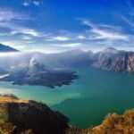 Panoramic view of a caldera lake with turquoise water, surrounded by rugged mountains and a blue sky.