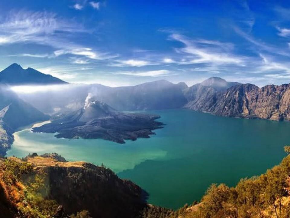 Panoramic view of a caldera lake with turquoise water, surrounded by rugged mountains and a blue sky.