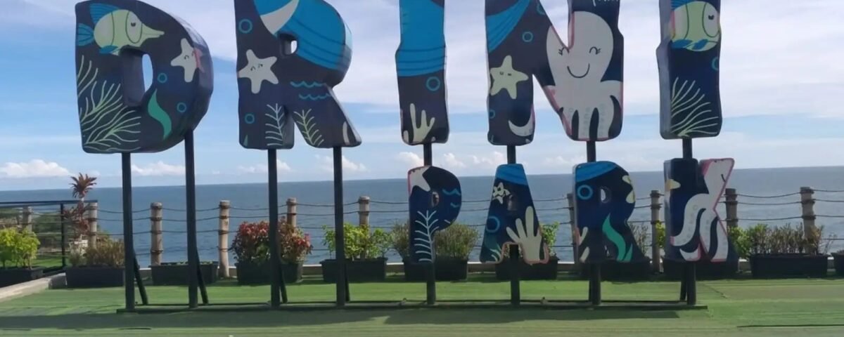 Large decorative letters with ocean-themed artwork mounted on posts at a seaside park, spelling a word or name beside the ocean view.