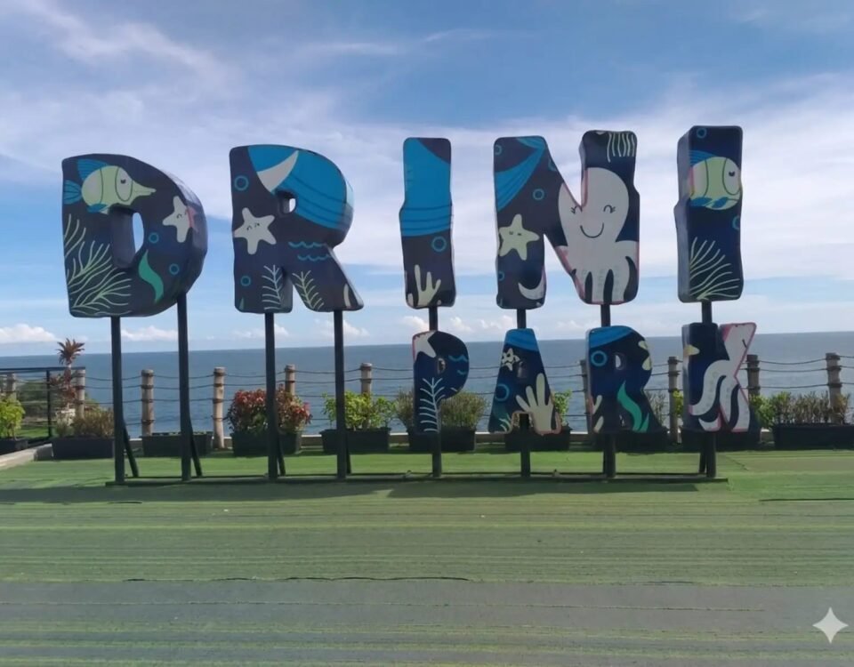 Large decorative letters with ocean-themed artwork mounted on posts at a seaside park, spelling a word or name beside the ocean view.