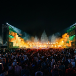 Nighttime outdoor concert with a large crowd, a brightly lit stage with orange and green lighting, and illuminated temple ruins in the background
