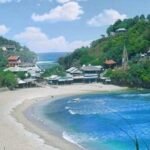 Tropical bay with a crescent sandy beach, turquoise water, and a hillside village of cottages amid green trees and blue sky.
