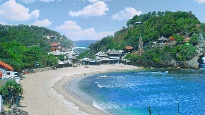 Tropical bay with a crescent sandy beach, turquoise water, and a hillside village of cottages amid green trees and blue sky.