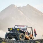 Off-road jeep parked on rocky slope with a large mountain in the background; safety flagging gear visible on the vehicle edge.