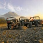 Three off-road jeeps parked on a rocky hillside with a distant mountain at sunset.