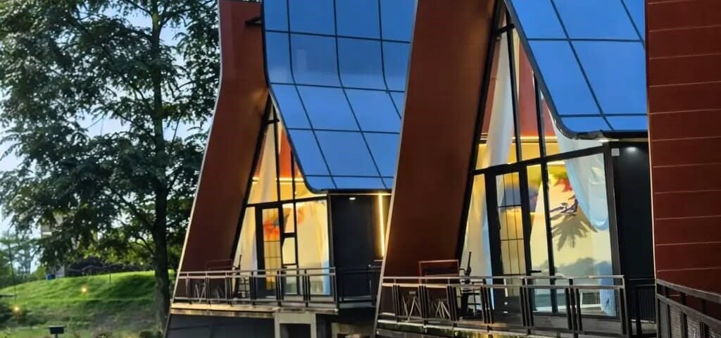 Modern building with tall blue glass panels and red-brown supports, outdoor walkway, and seating area with red benches at dusk.