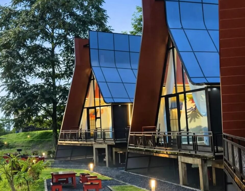 Modern building with tall blue glass panels and red-brown supports, outdoor walkway, and seating area with red benches at dusk.