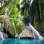 Turquoise pool fed by moss-covered waterfalls in a lush green forest on a sunny day.