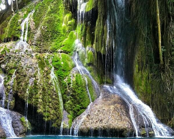 Turquoise pool fed by moss-covered waterfalls in a lush green forest on a sunny day.