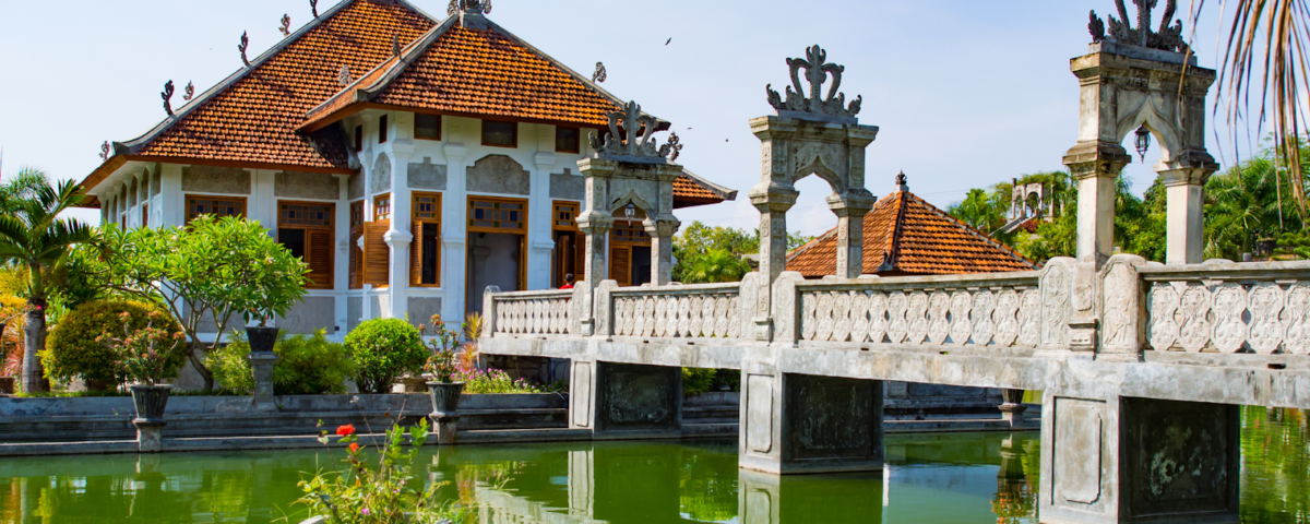 Balinese-style courtyard with a red-tiled pavilion and an ornate stone bridge over a green reflecting pool surrounded by plants