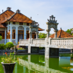 Balinese-style courtyard with a red-tiled pavilion and an ornate stone bridge over a green reflecting pool surrounded by plants