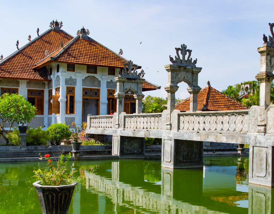 Balinese-style courtyard with a red-tiled pavilion and an ornate stone bridge over a green reflecting pool surrounded by plants