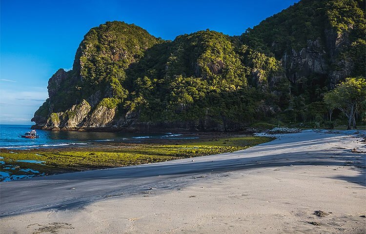 Tropical beach with white sand, calm blue water, and a forested rocky cliff; a small boat near the shore on the left.
