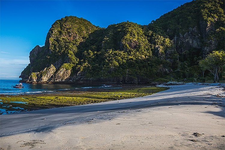 Tropical beach with white sand, calm blue water, and a forested rocky cliff; a small boat near the shore on the left.