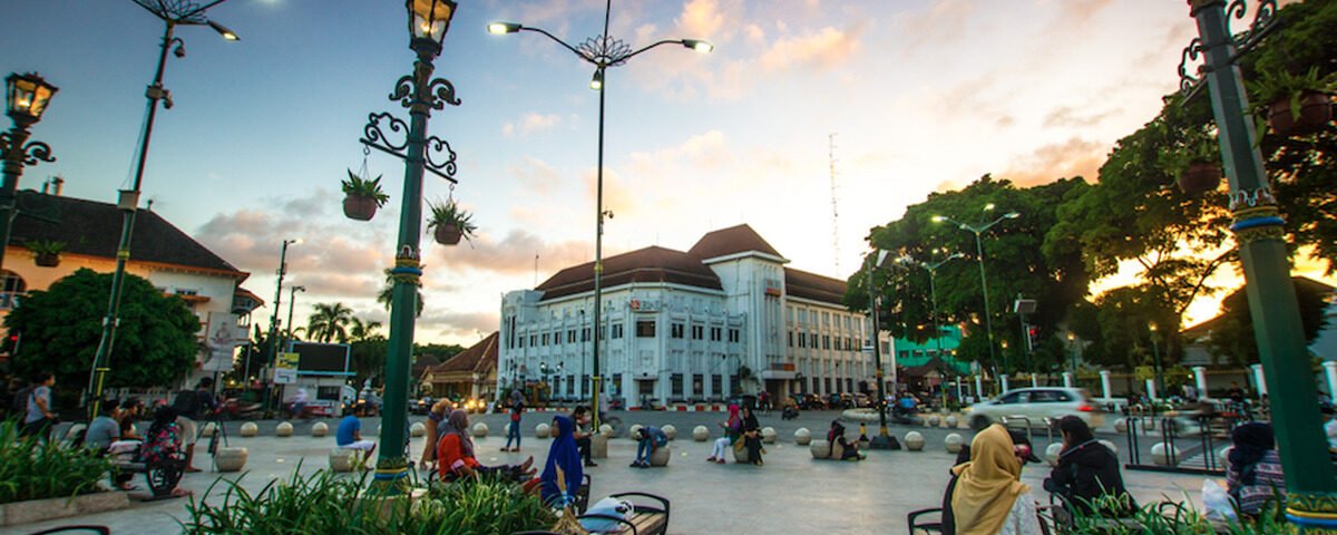 Public square with ornate street lamps, benches, and people sitting near a white colonial-style building at sunset.