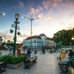 Public square with ornate street lamps, benches, and people sitting near a white colonial-style building at sunset.