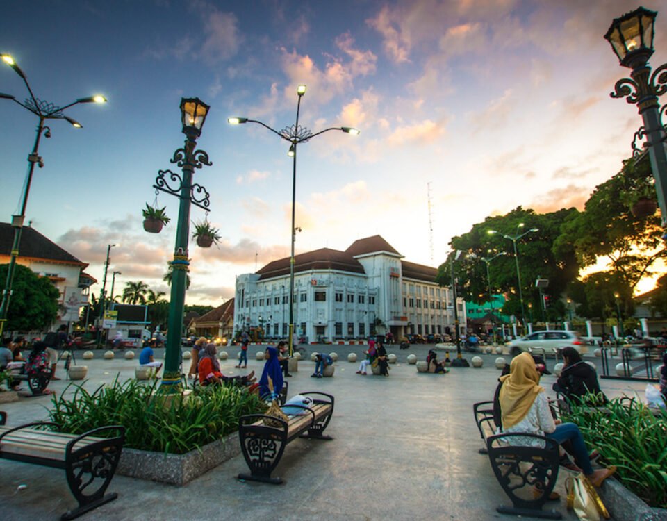 Public square with ornate street lamps, benches, and people sitting near a white colonial-style building at sunset.