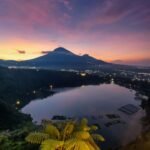 Sunset over a volcanic mountain with a lake in the foreground and city lights along the shore.