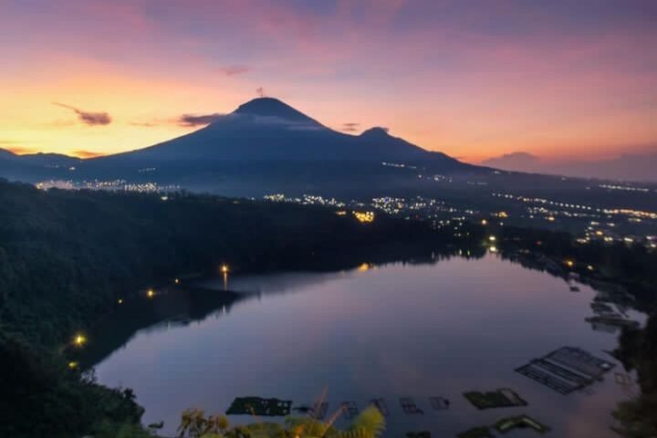 Sunset over a volcanic mountain with a lake in the foreground and city lights along the shore.