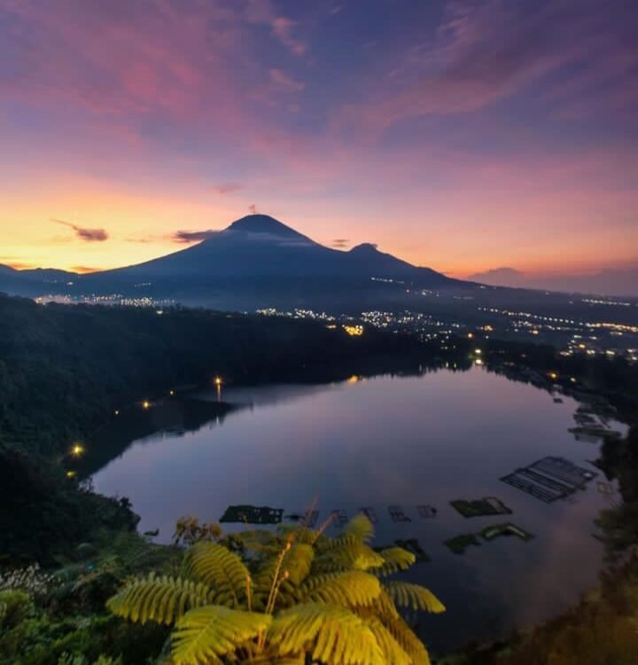 Sunset over a volcanic mountain with a lake in the foreground and city lights along the shore.