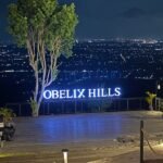 Night view from Obelix Hills terrace with a glowing 'OBELIX HILLS' sign, a tall tree, and a city of lights in the distance.