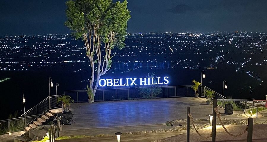 Night view from Obelix Hills terrace with a glowing 'OBELIX HILLS' sign, a tall tree, and a city of lights in the distance.