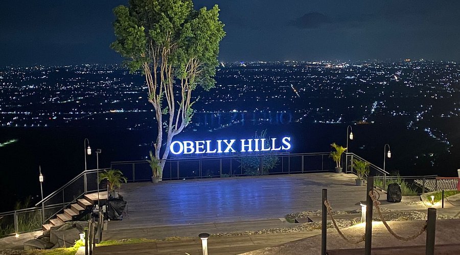 Night view from Obelix Hills terrace with a glowing 'OBELIX HILLS' sign, a tall tree, and a city of lights in the distance.