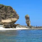 Coastal scene with a large rock formation on the left, sea stack in the distance, and a sandy shoreline under a clear blue sky.