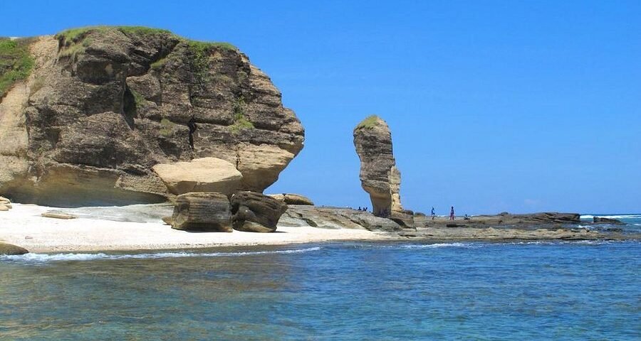 Coastal scene with a large rock formation on the left, sea stack in the distance, and a sandy shoreline under a clear blue sky.