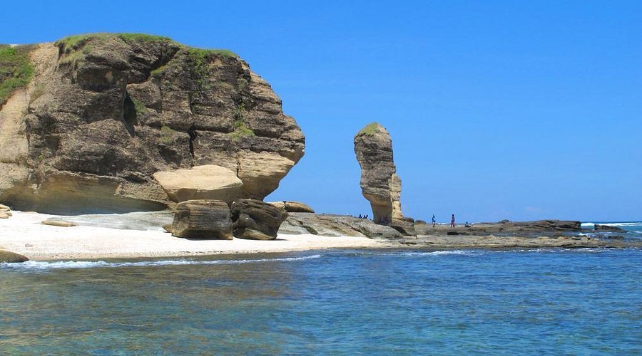 Coastal scene with a large rock formation on the left, sea stack in the distance, and a sandy shoreline under a clear blue sky.