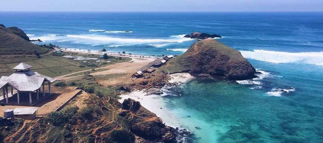 A coastal landscape with turquoise water, rocky promontory, and an open-air pavilion on the left overlooking the sea.