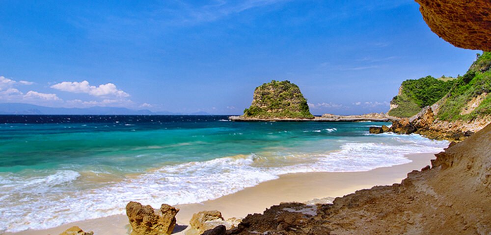 Sunny beach with clear turquoise water, white-capped waves, and rocky cliffs on the right under a bright blue sky.