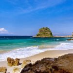 Sunny beach with clear turquoise water, white-capped waves, and rocky cliffs on the right under a bright blue sky.