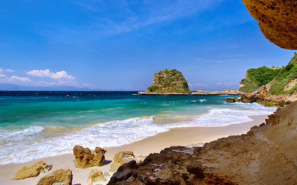 Sunny beach with clear turquoise water, white-capped waves, and rocky cliffs on the right under a bright blue sky.