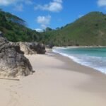 Sunny tropical beach with pale sand, rocky outcrops in the foreground, turquoise water, and green hills in the distance.