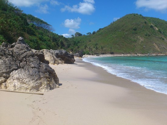 Sunny tropical beach with pale sand, rocky outcrops in the foreground, turquoise water, and green hills in the distance.