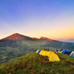Campsite on a grassy hill at sunset with colorful tents facing a distant volcanic crater and ridge line.