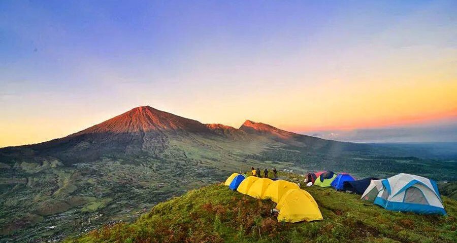 Campsite on a grassy hill at sunset with colorful tents facing a distant volcanic crater and ridge line.